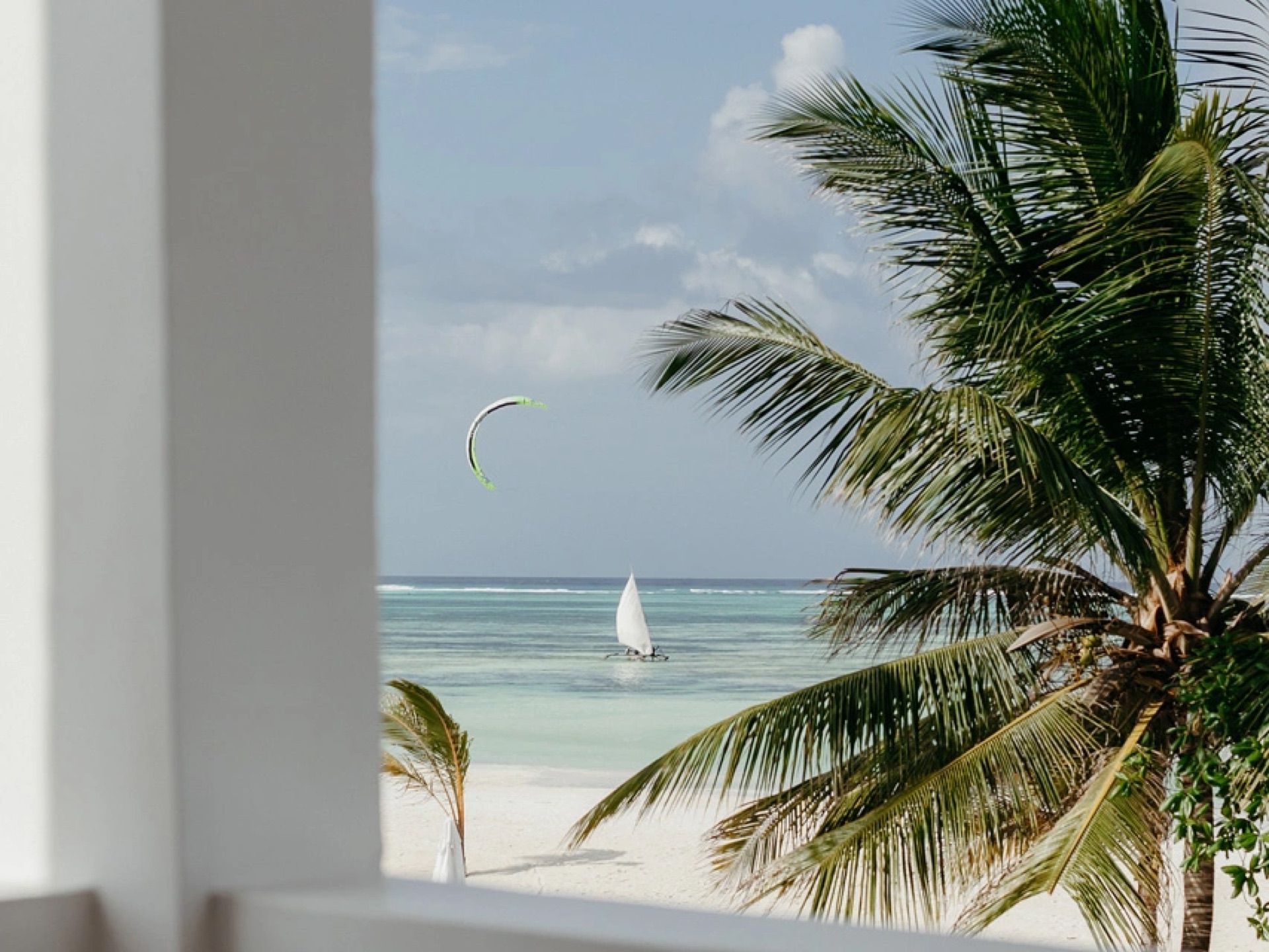 A view past a whitewashed wall to white sand, turquoise water, a dhow under sail and a kitesurfing kite